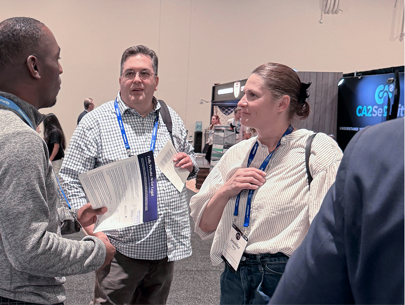 Three people engaged in conversation at a conference or event, holding papers and wearing lanyards with badges.