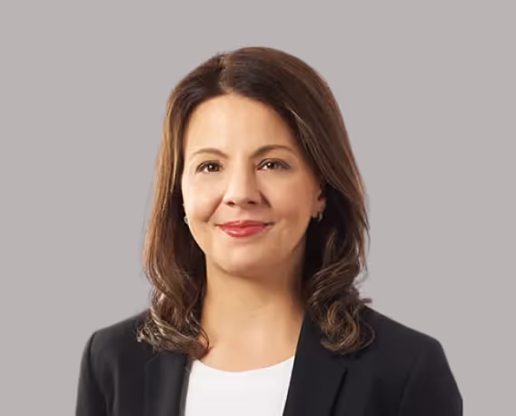 Professional woman with shoulder-length brown hair wearing a black blazer and white top, smiling against a gray background.