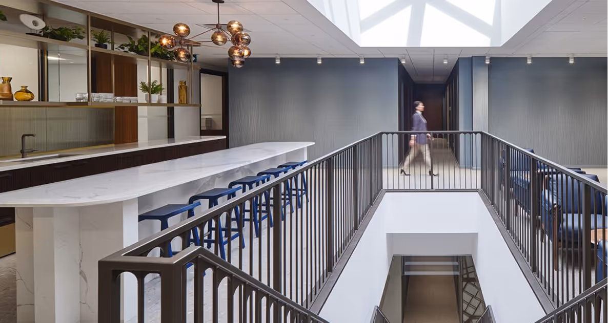Modern office interior with a long white marble counter, blue stools, pendant lighting, and a woman walking by a railing overlooking a staircase.