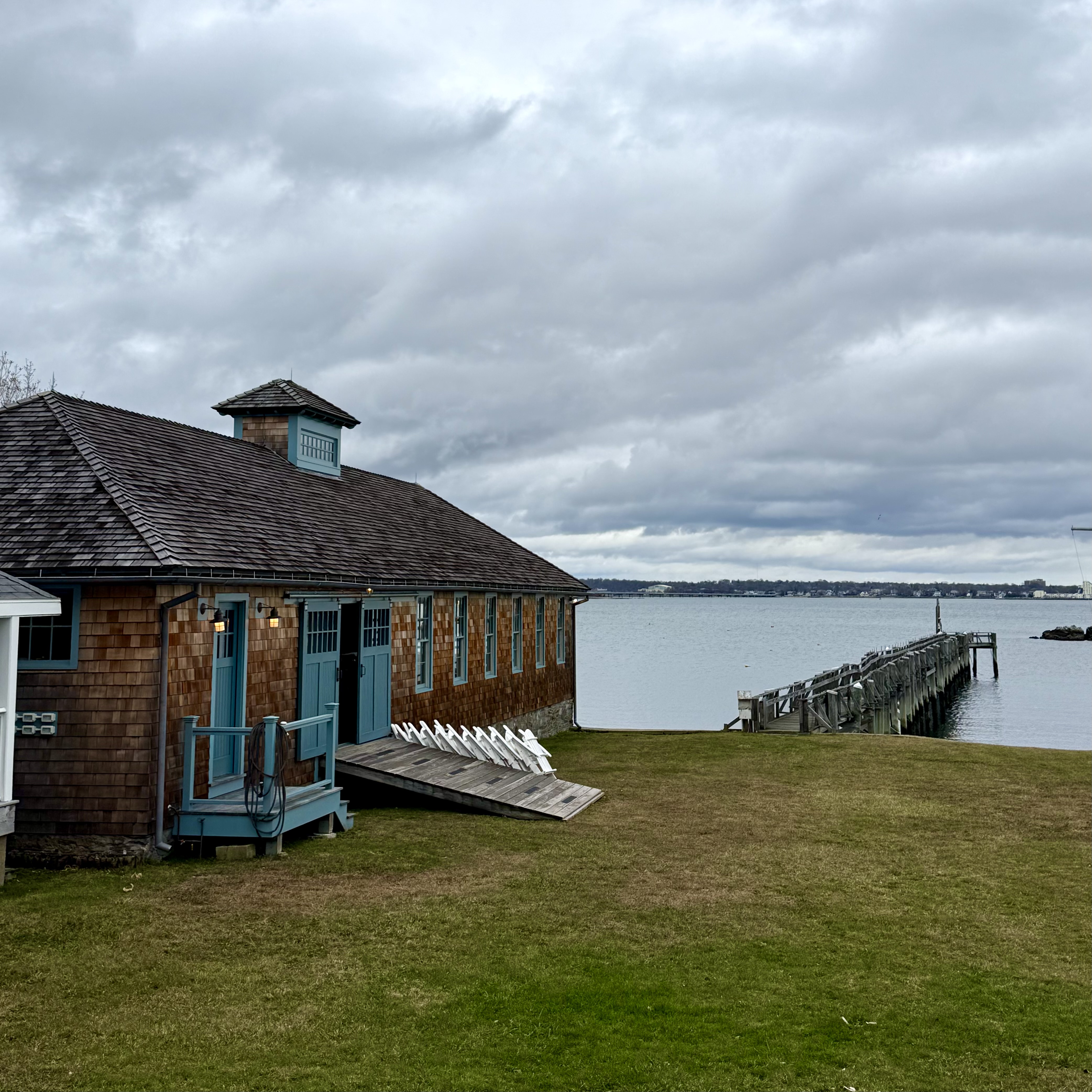 A shingled boathouse on the shore under a grey sky