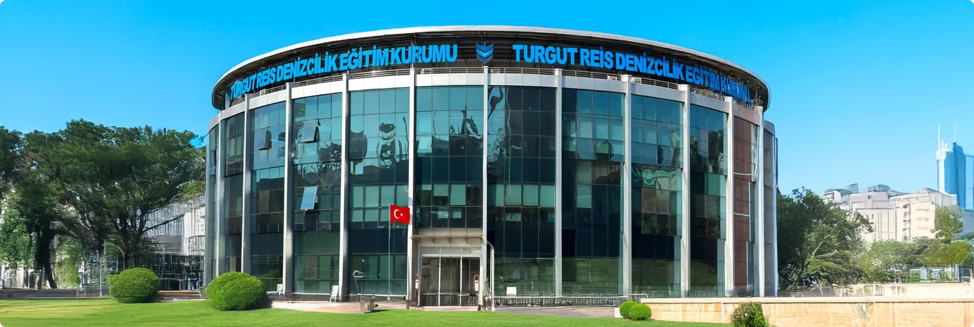 Turgut Reis Denizcilik Eğitim Kurumu yazısı ve Türk bayrağı bulunan modern cam cepheli yuvarlak bina.