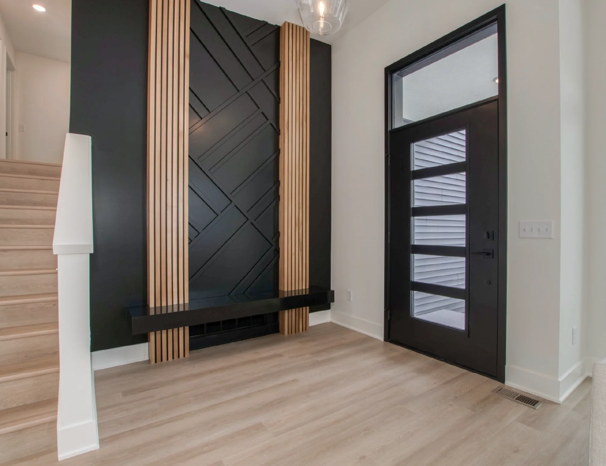 Modern entryway with light wood floors, black front door with horizontal glass panels, and a black wall accent with vertical wooden slats next to stairs.