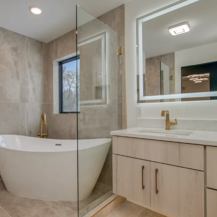 Modern bathroom with a freestanding white bathtub, beige tiled walls, gold faucet, glass shower partition, and a vanity with illuminated mirror and gold fixtures.