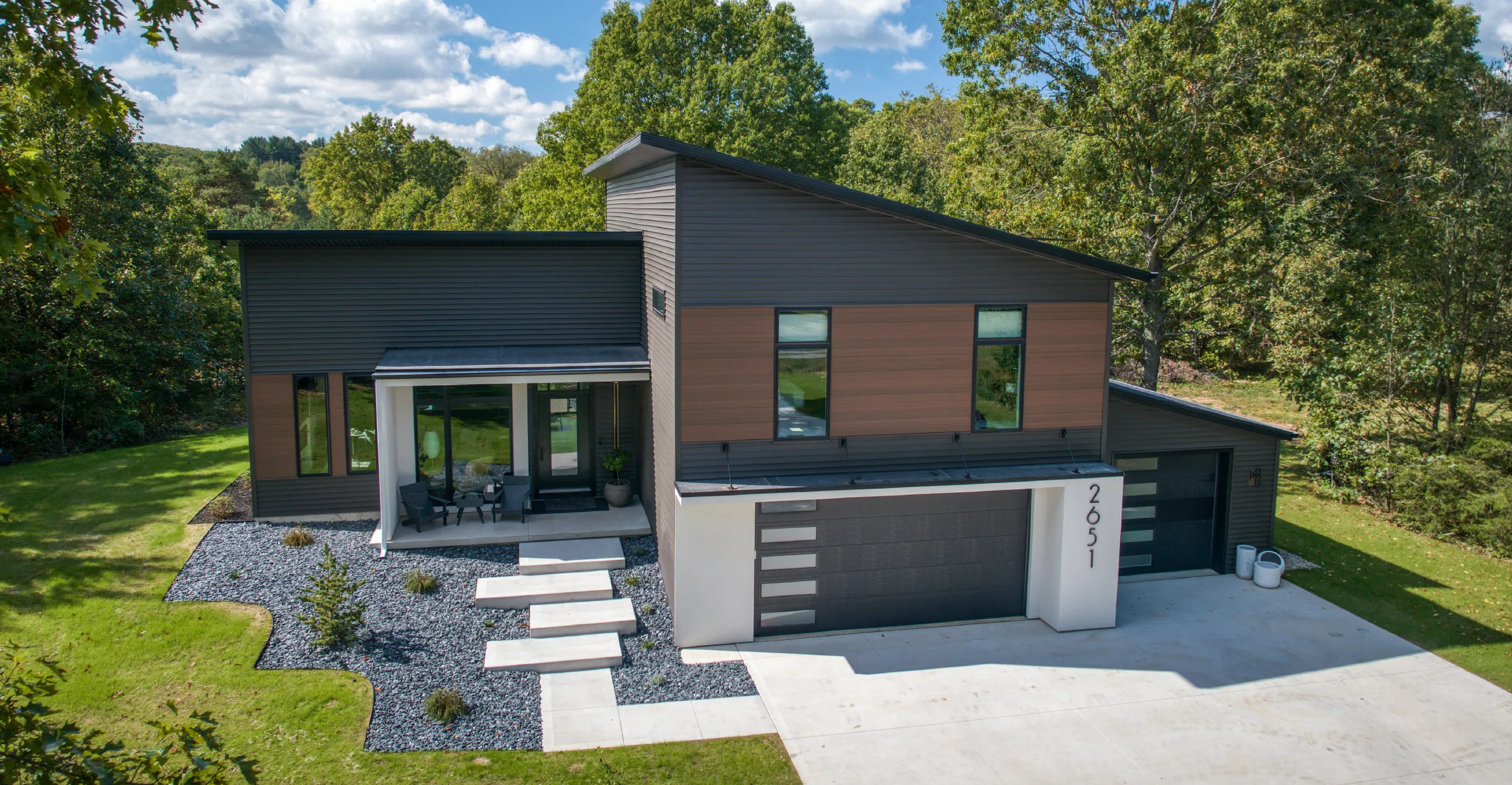 Modern two-story house with gray and brown exterior, large windows, concrete driveway, and a small front porch with chairs.