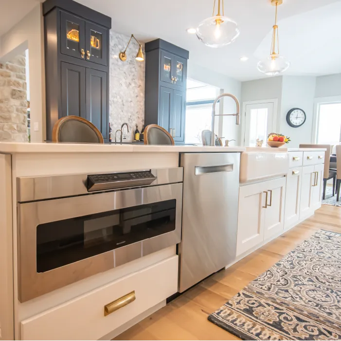 Modern kitchen with white cabinetry, stainless steel microwave and dishwasher, farmhouse sink, dark wood cabinets, and a patterned rug on light wood floor.