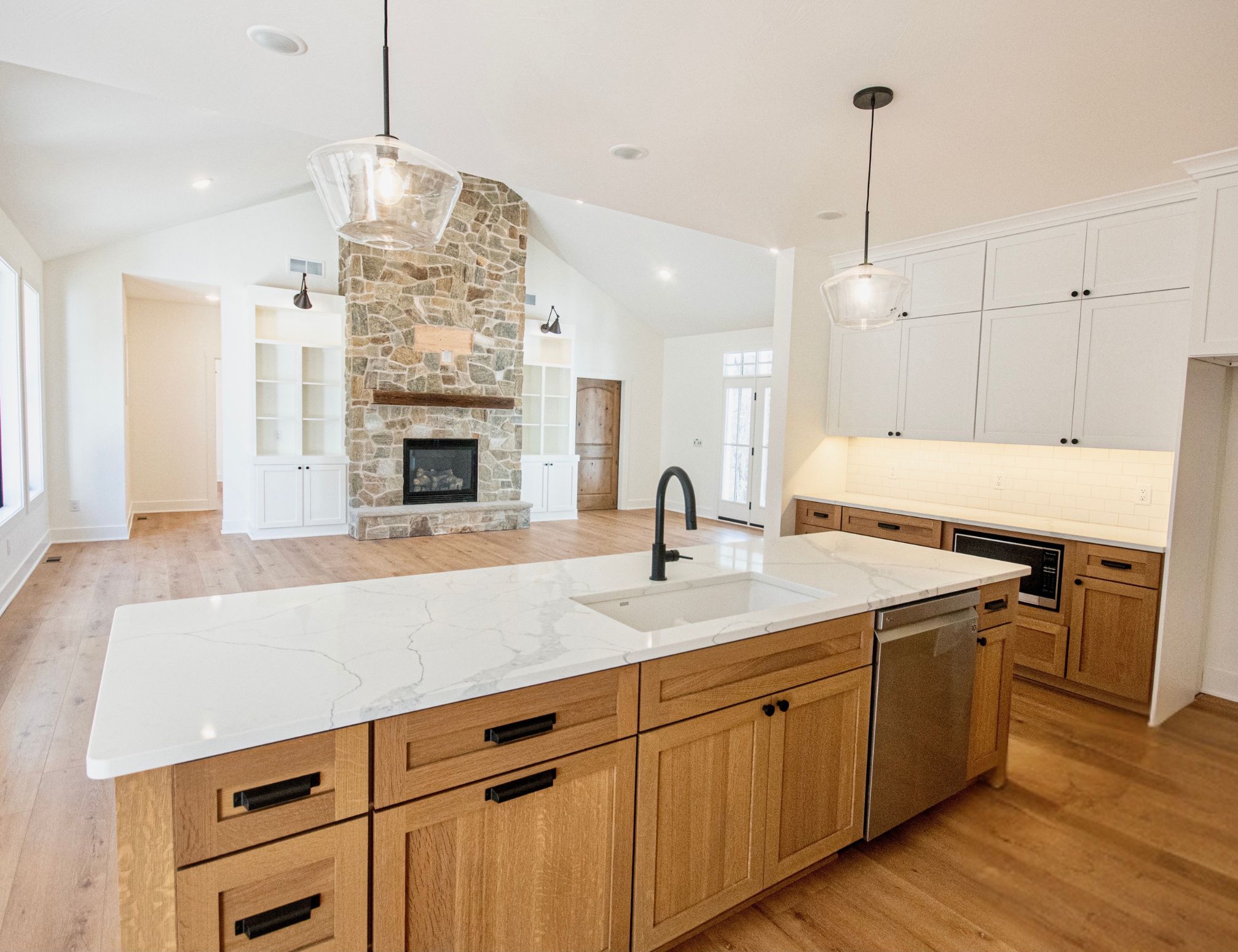 Modern open kitchen with a large marble countertop island, wooden cabinets, pendant lights, and a stone fireplace in the adjacent living area.