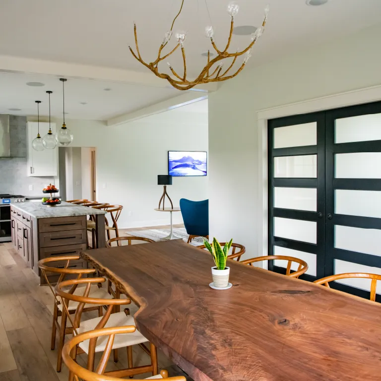 Modern dining room with a large wooden table surrounded by wooden chairs, a potted plant centerpiece, and a kitchen with island and pendant lights in the background.