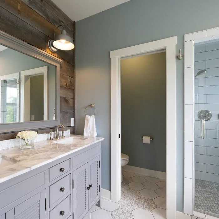 Modern bathroom with a gray vanity topped with marble, a large mirror, wall-mounted light, and a doorway leading to a toilet room with hexagonal patterned floor tiles.
