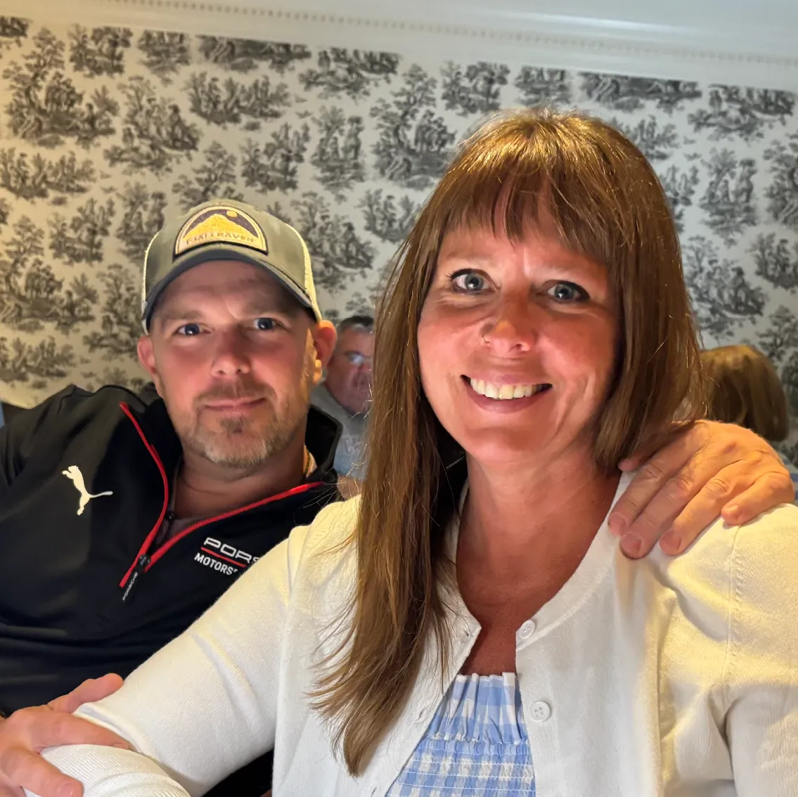 Smiling woman in a white cardigan with a man wearing a cap and a Porsche jacket behind her with his hand on her shoulder.