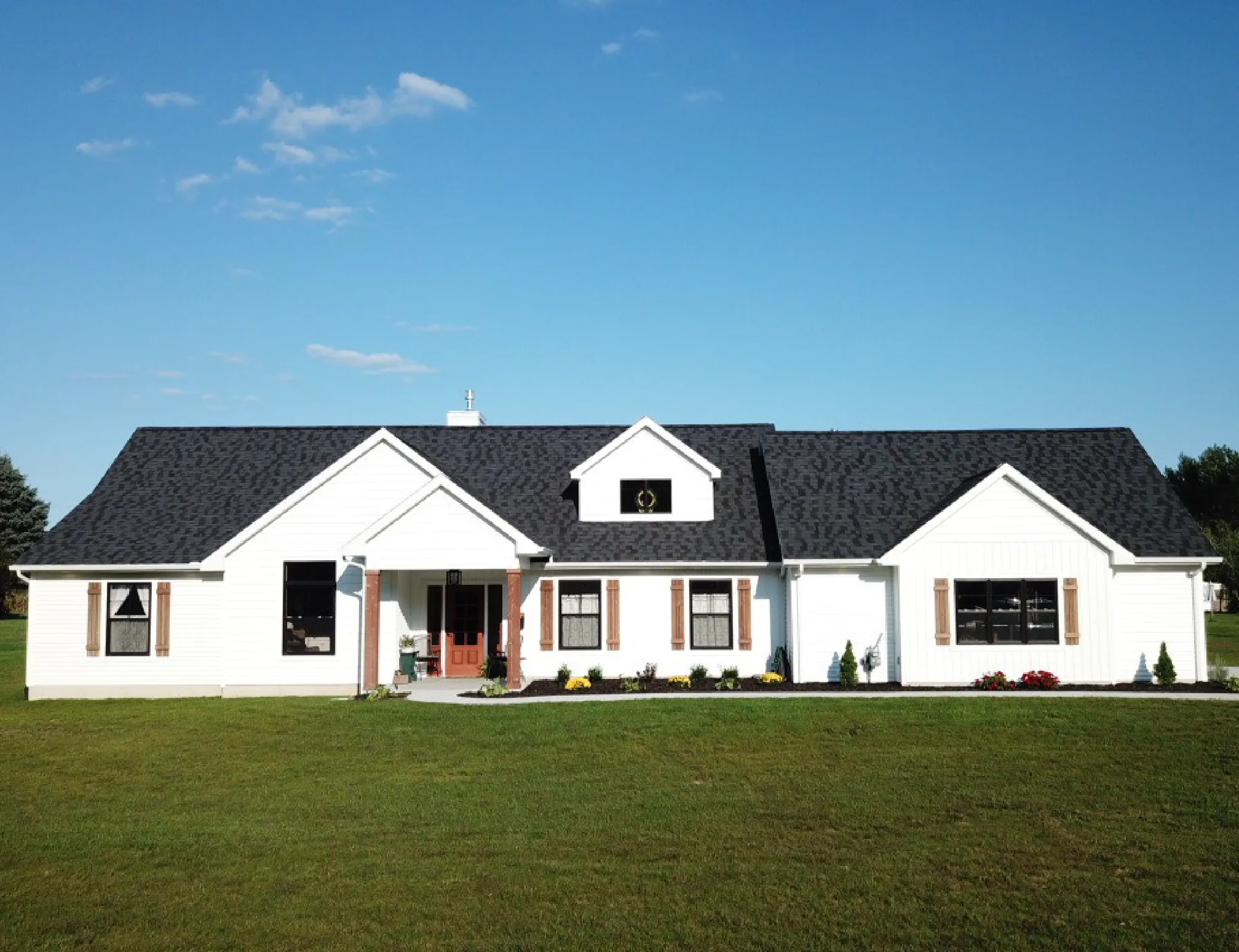 Single-story white house with dark roof, wooden shutters, a red front door, and a green lawn under a blue sky.
