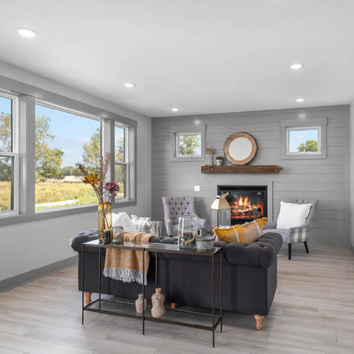 Cozy living room with a dark sofa, two armchairs, a lit fireplace, and large windows showing a sunny outdoor view.