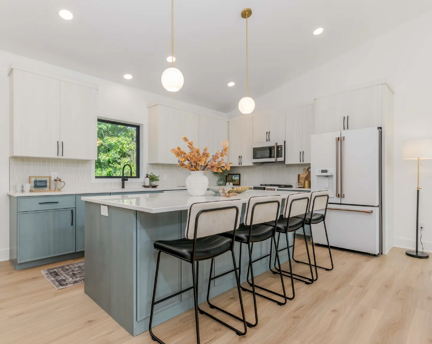 Modern kitchen with light blue and white cabinets, three black and white bar stools at a large island, and pendant lights overhead.
