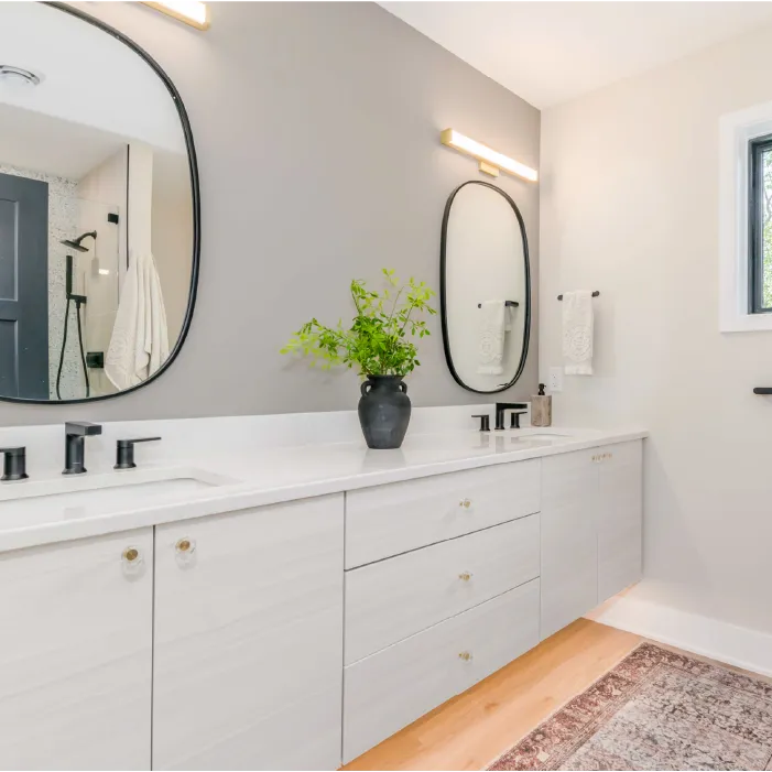 Modern bathroom with white double vanity, black oval mirrors, black faucets, a black vase with green plant, and a light wood floor with a patterned rug.