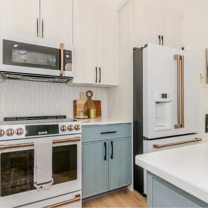 Modern kitchen corner with white and blue cabinets, white stove with copper knobs, matching microwave, and a white refrigerator with copper handles.