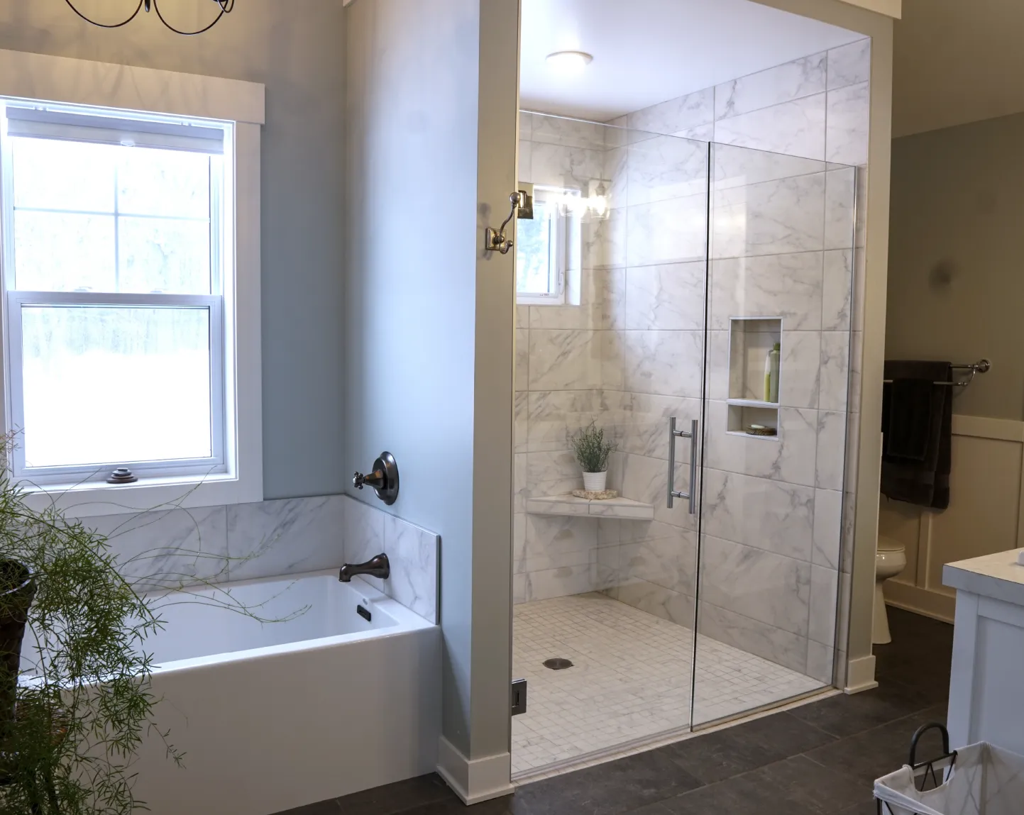 Modern bathroom with a white bathtub under a window, a glass-enclosed marble-tiled shower with built-in shelf and bench, and dark floor tiles.