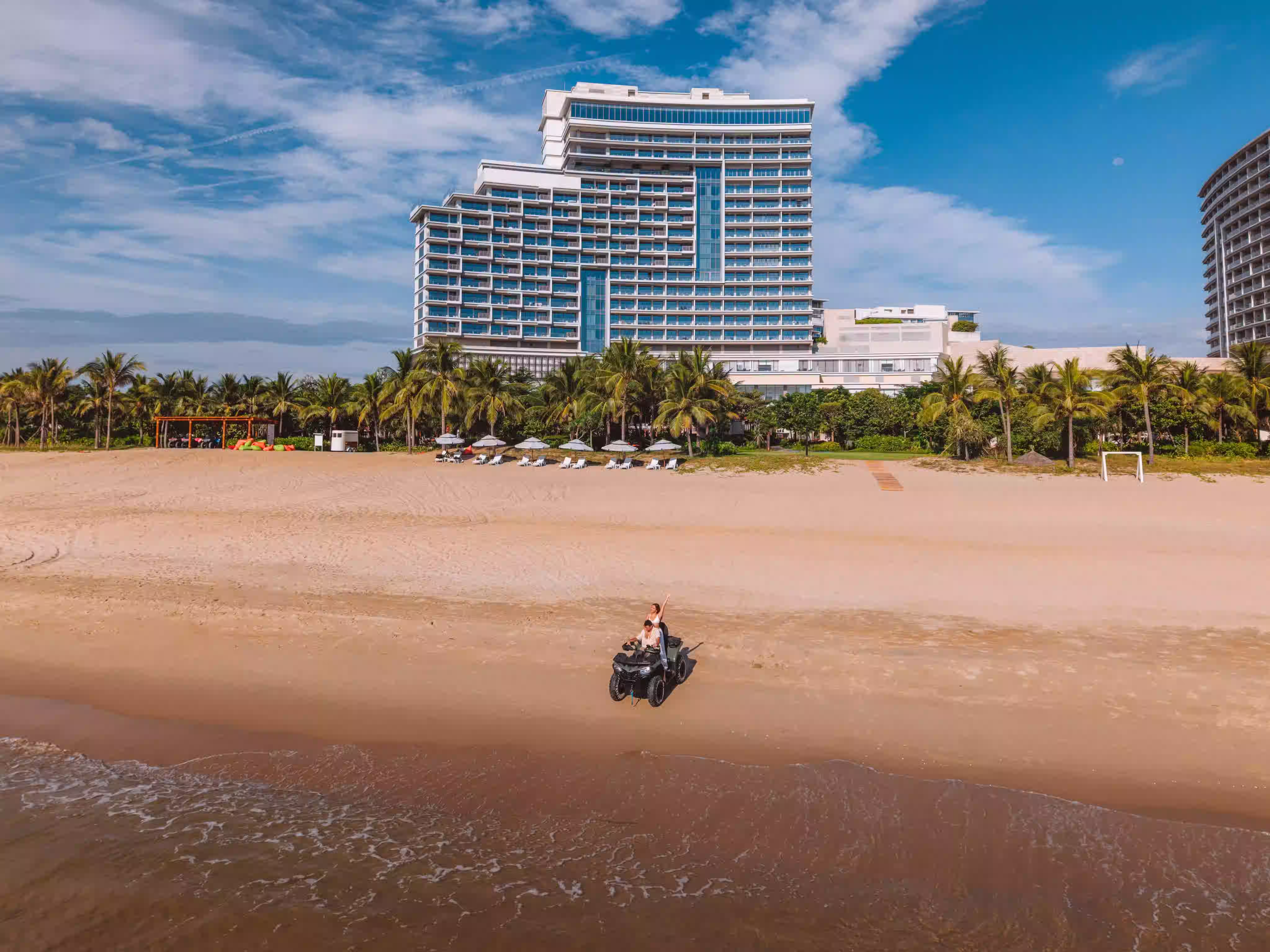 Guests enjoying an ATV ride along the beach at Hoiana Hotel & Suites, with the palm-lined property stretching behind them.