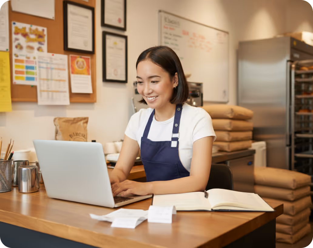 Woman wearing apron smiling while working on a laptop at a wooden counter with receipts and an open notebook.