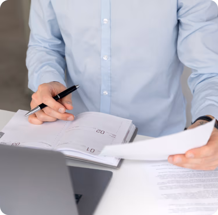 Person in a light blue shirt holding a pen and reviewing an open planner and documents on a white desk with a laptop.