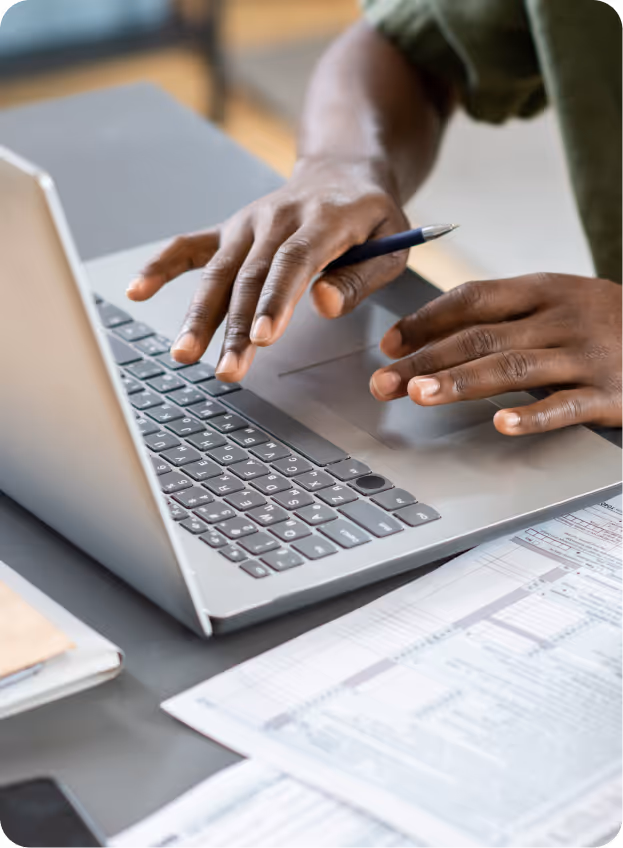 Person typing on a laptop keyboard with a pen in one hand, working at a desk with documents and papers.