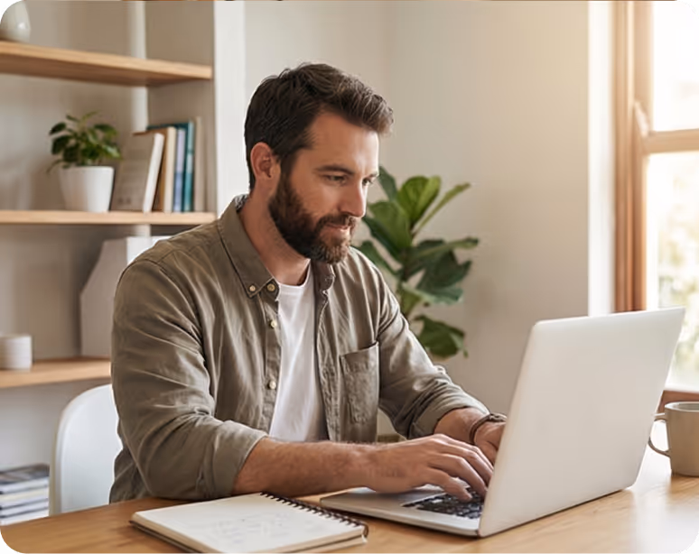 Man with beard typing on a laptop at a wooden table with a notebook and coffee mug nearby.