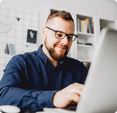 Man with glasses and beard smiling while working on a laptop in a home office.