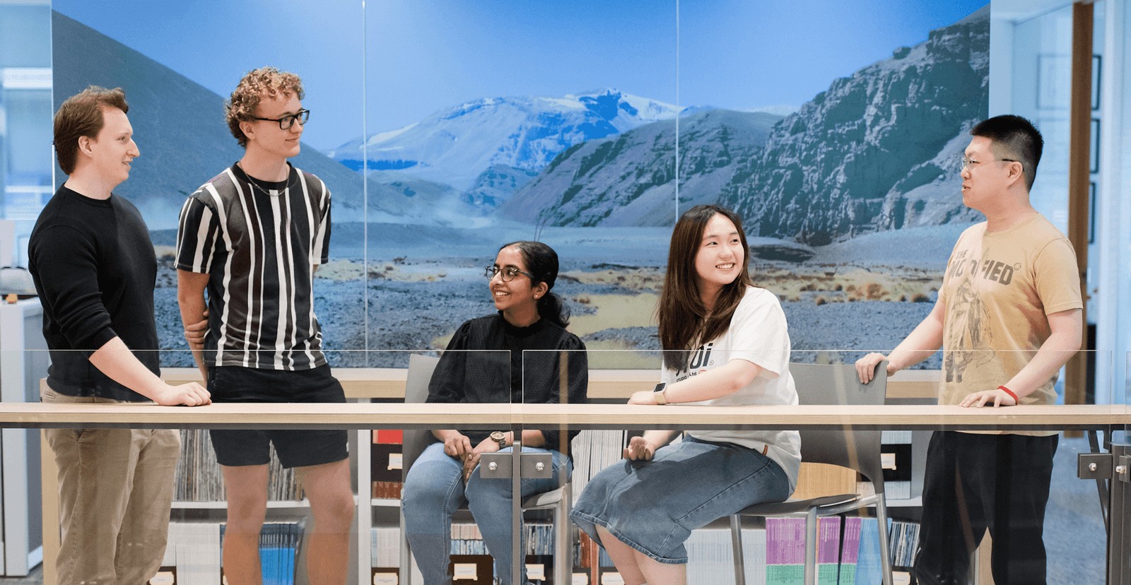 Five people casually talking and smiling in a modern office with a mountain landscape mural behind them.