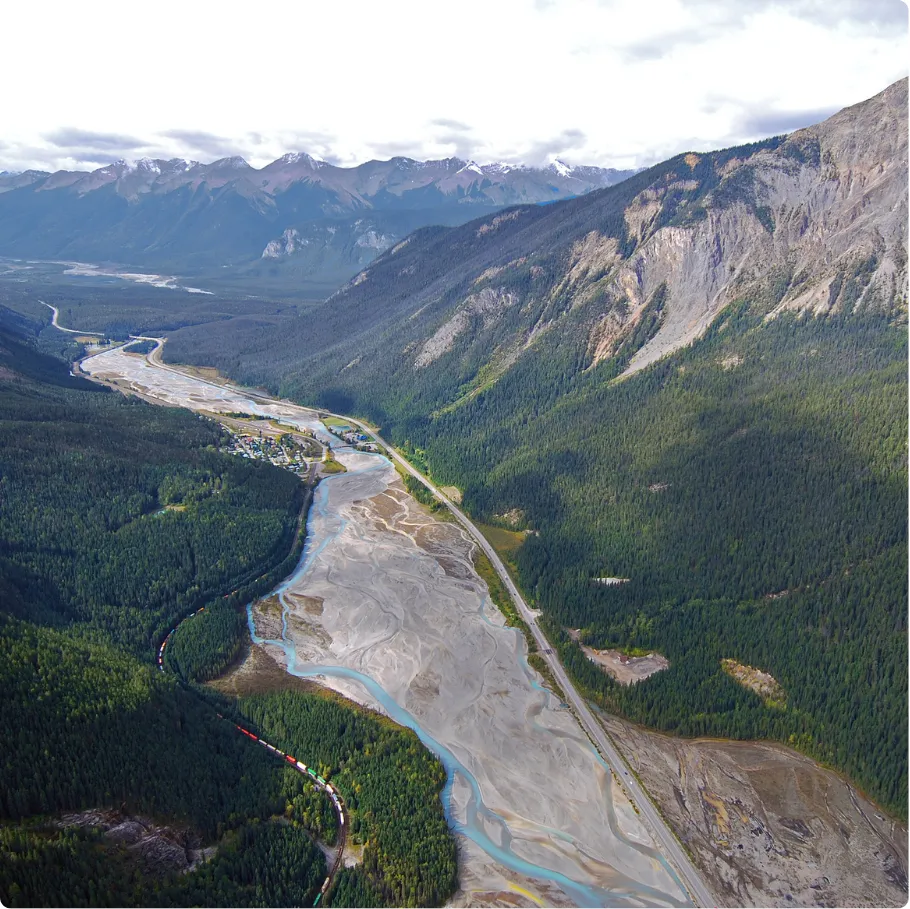 Aerial view of a winding river with light blue water flowing through a wide, sandy riverbed surrounded by dense green forest and mountains with rugged rocky peaks under a cloudy sky.