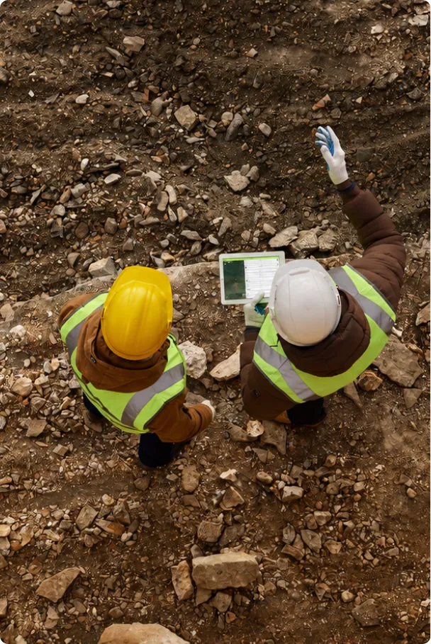 Overhead view of two construction workers in reflective vests and helmets reviewing data on a tablet while standing on rocky soil at a construction site.
