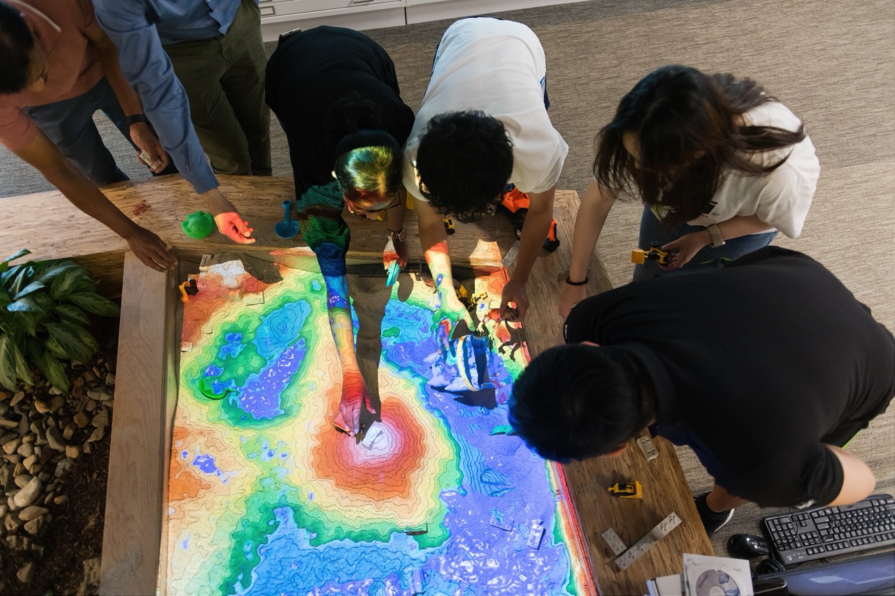 Five people gathered around an interactive sandbox table with topographic projections in colors like blue, green, and red, manipulating the sand and using small tools.