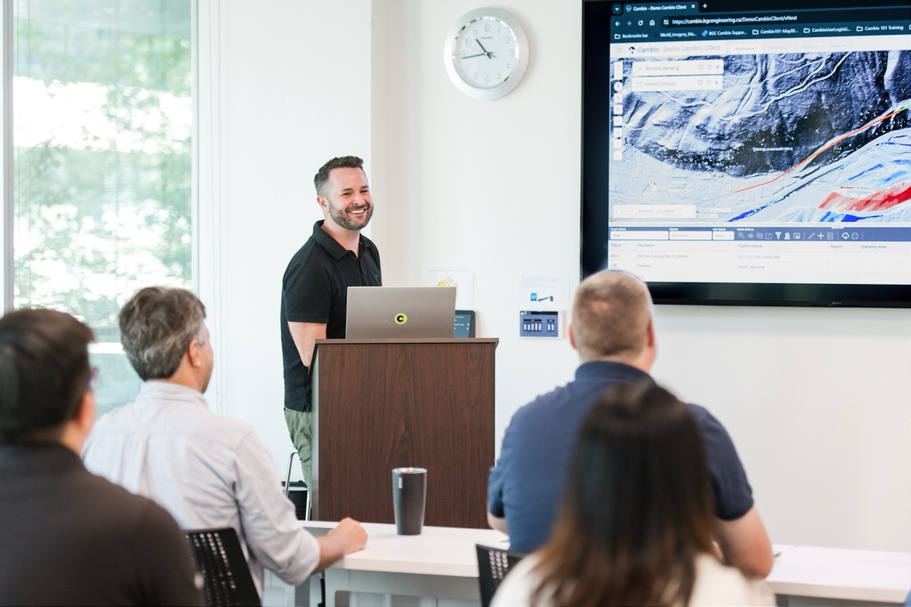 A man in a black polo shirt stands at a lectern smiling while presenting to a small audience seated at tables, with a large screen on the wall displaying a topographic map and data.