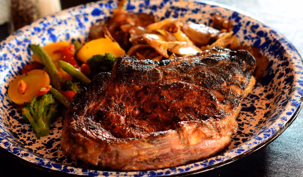 A large, grilled ribeye steak served with colorful sautéed vegetables and crispy potatoes on a blue-speckled plate. 