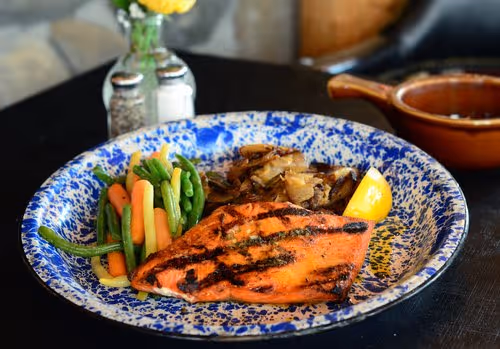 A grilled salmon fillet garnished with lemon, served alongside sautéed vegetables and seasoned potatoes on a decorative plate.