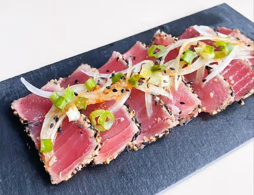 Sliced seared tuna served on a black slate plate, topped with green onions and white onion, showcasing a vibrant pink color.
