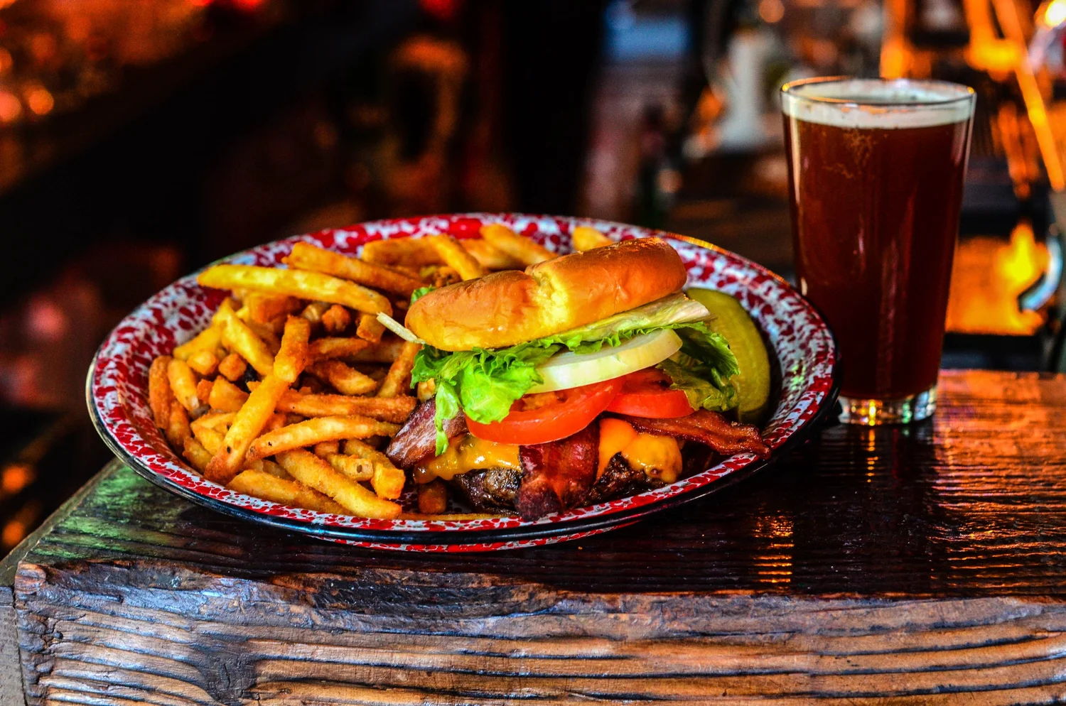 A juicy bacon cheeseburger topped with lettuce, tomato, and onions, served with golden fries and a pint of beer on a rustic table.