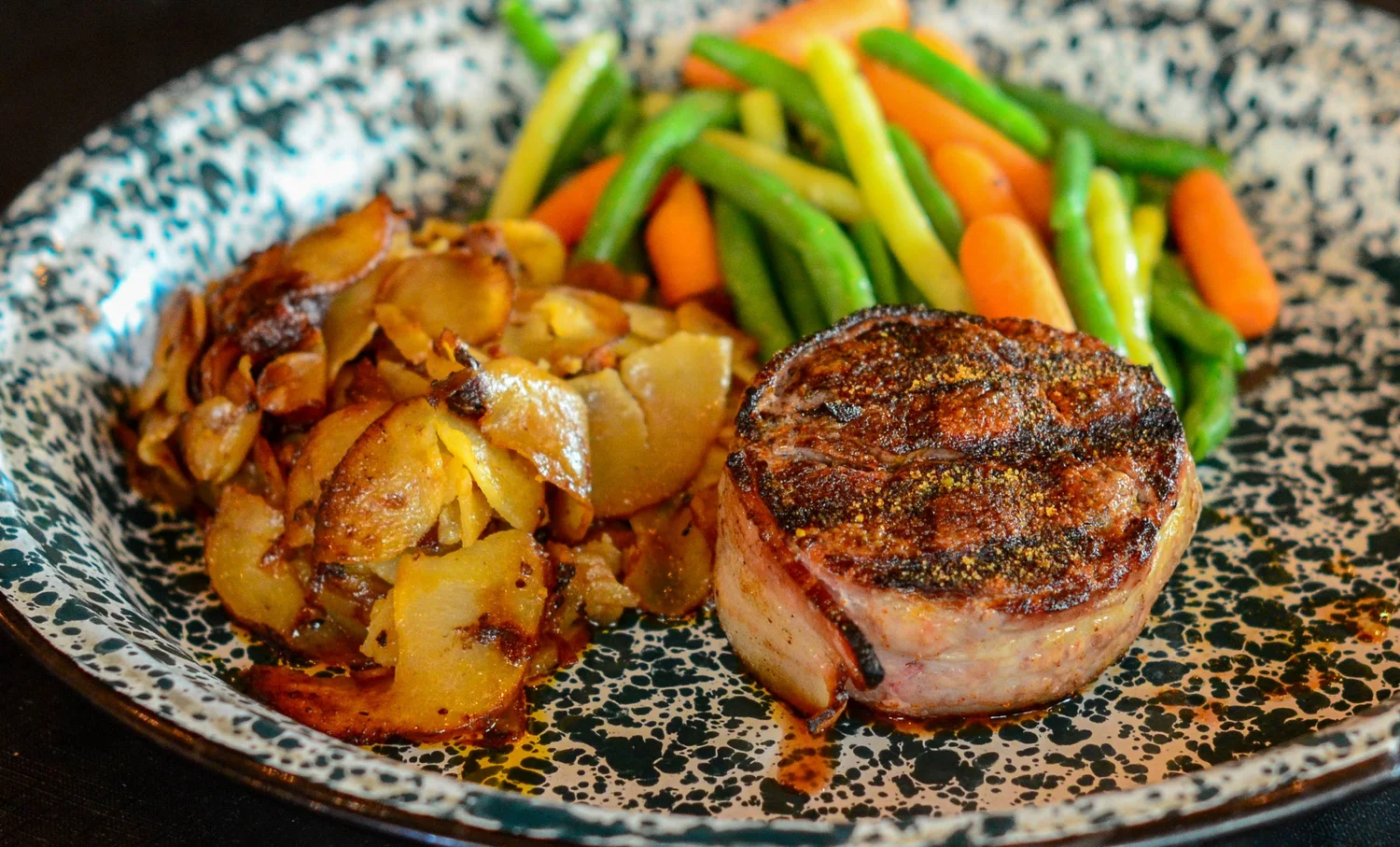 A plate featuring a grilled beef fillet, crispy sliced potatoes, and a side of vibrant green beans and carrots.