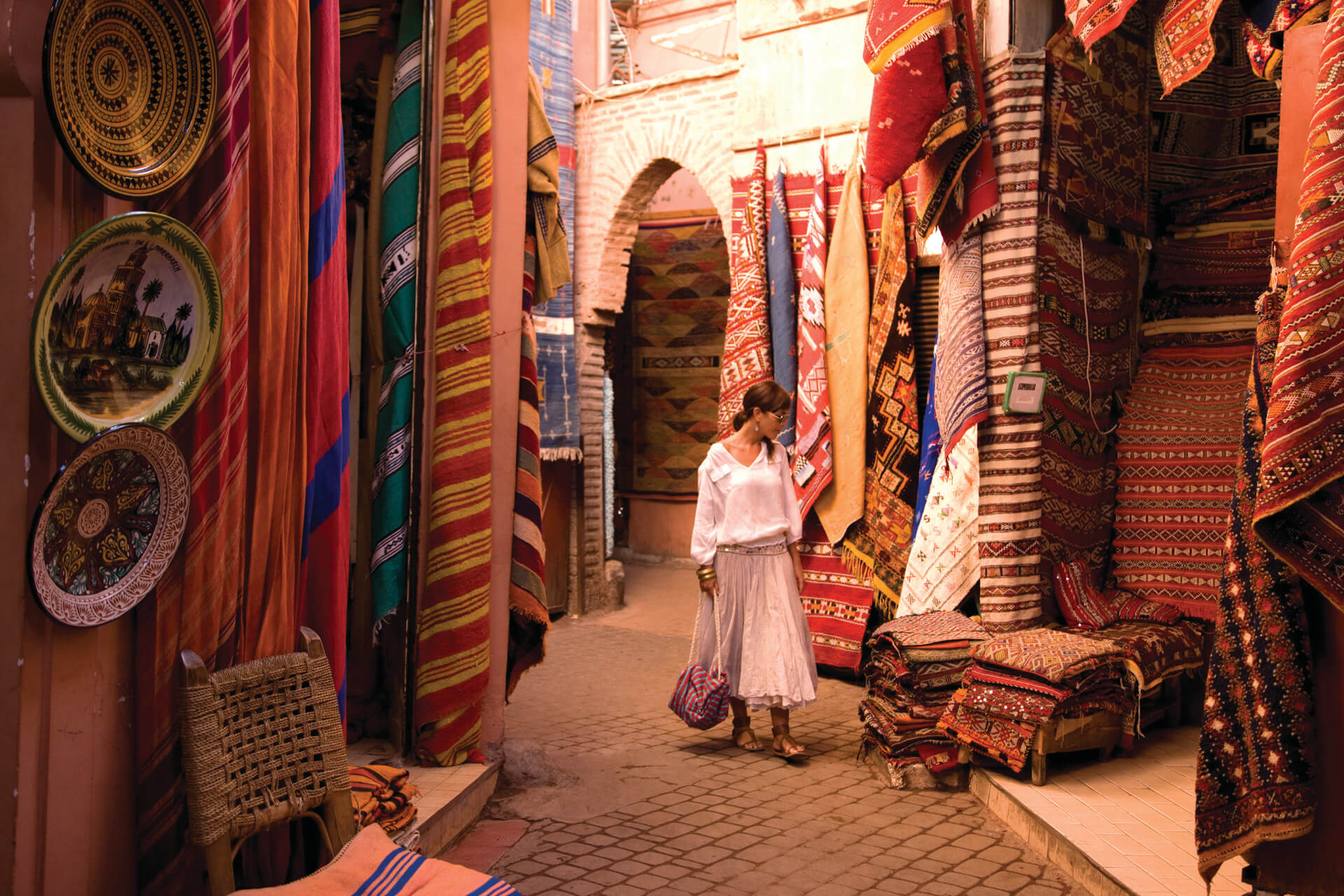 Woman in white blouse and skirt browsing patterned rugs and textiles in a narrow market alley in the medina in Marrakech with Moroccan Guides Travel during a day guided trip
