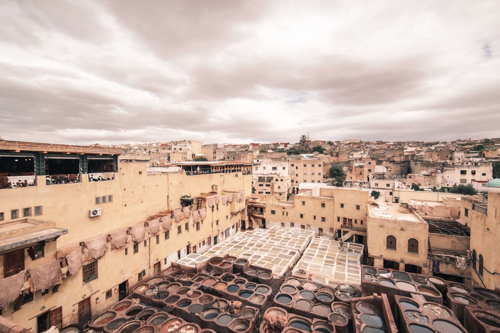 Traditional leather tannery in Fez morocco with large stone vats filled with various colored liquids, surrounded by beige buildings under a cloudy sky during a guided tour with Moroccan Guides Travel.