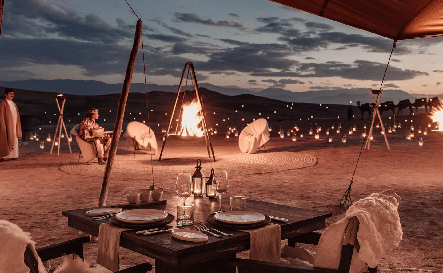 Agafay desert dinner table setting for two in the Agafay Desert camp at twilight, with a large central fire pit, glowing lanterns, and the Atlas Mountains visible in the background.