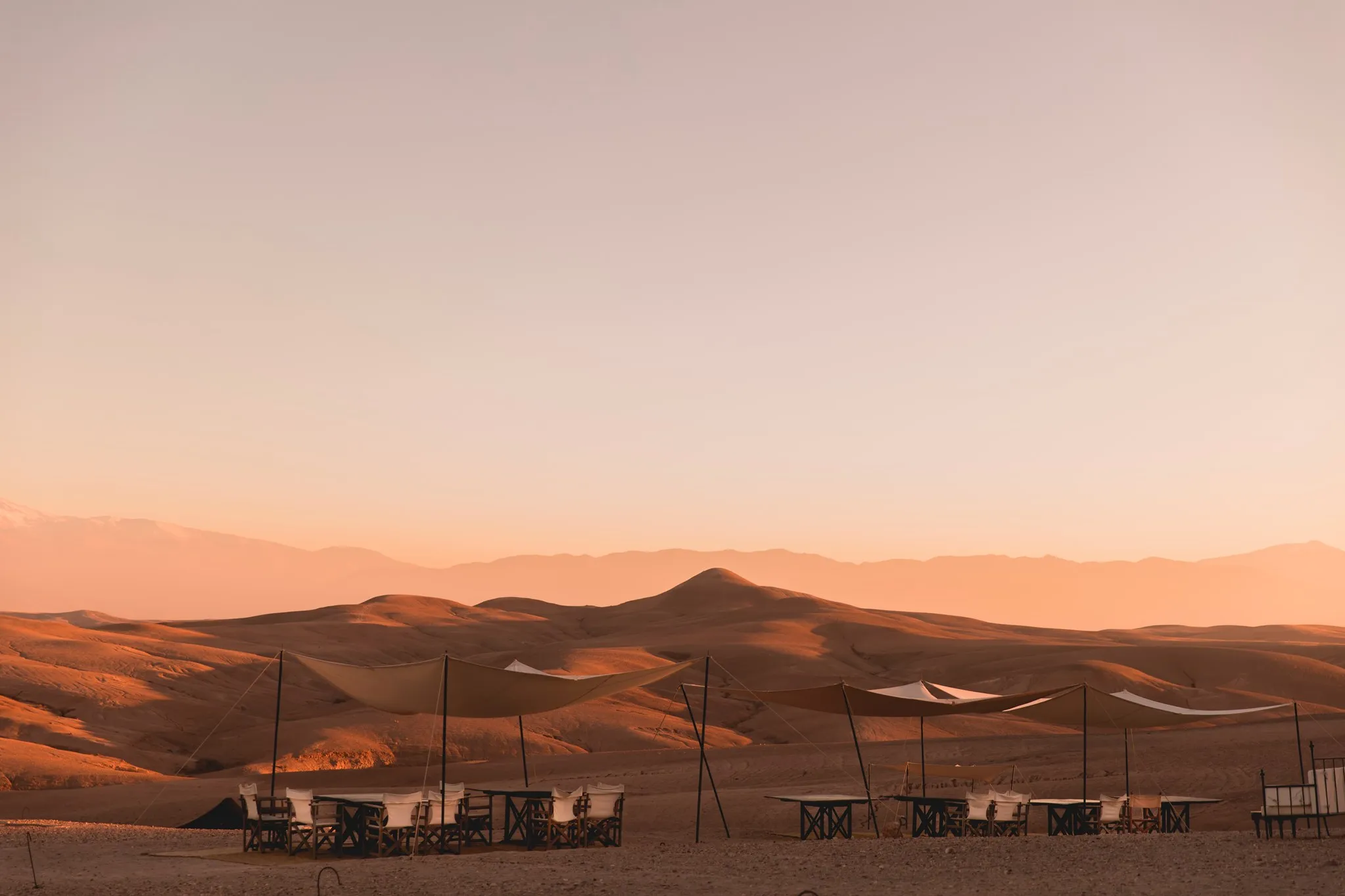 Panoramic view of the Agafay Desert at sunset, showing several dining tables protected by light canopies with the rolling brown hills and distant Atlas Mountains in the background with Moroccan Guides travel