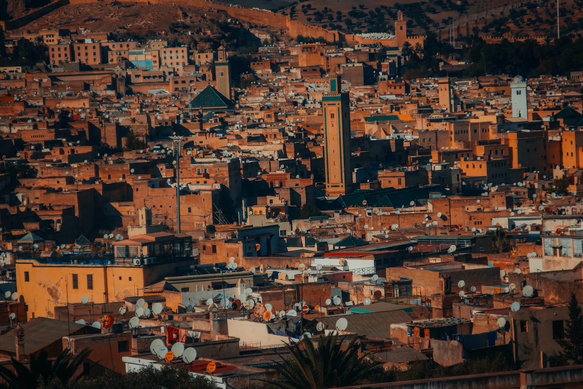 A panoramic view over the dense, historic cityscape of Fes, Morocco. The scene is filled with tightly packed, earth-toned buildings bathed in warm, golden sunlight. A tall, square minaret with intricate green tiling stands out among the low-rise structures. The rooftops are crowded with countless white satellite dishes, and a hillside featuring ancient city walls rises in the background.