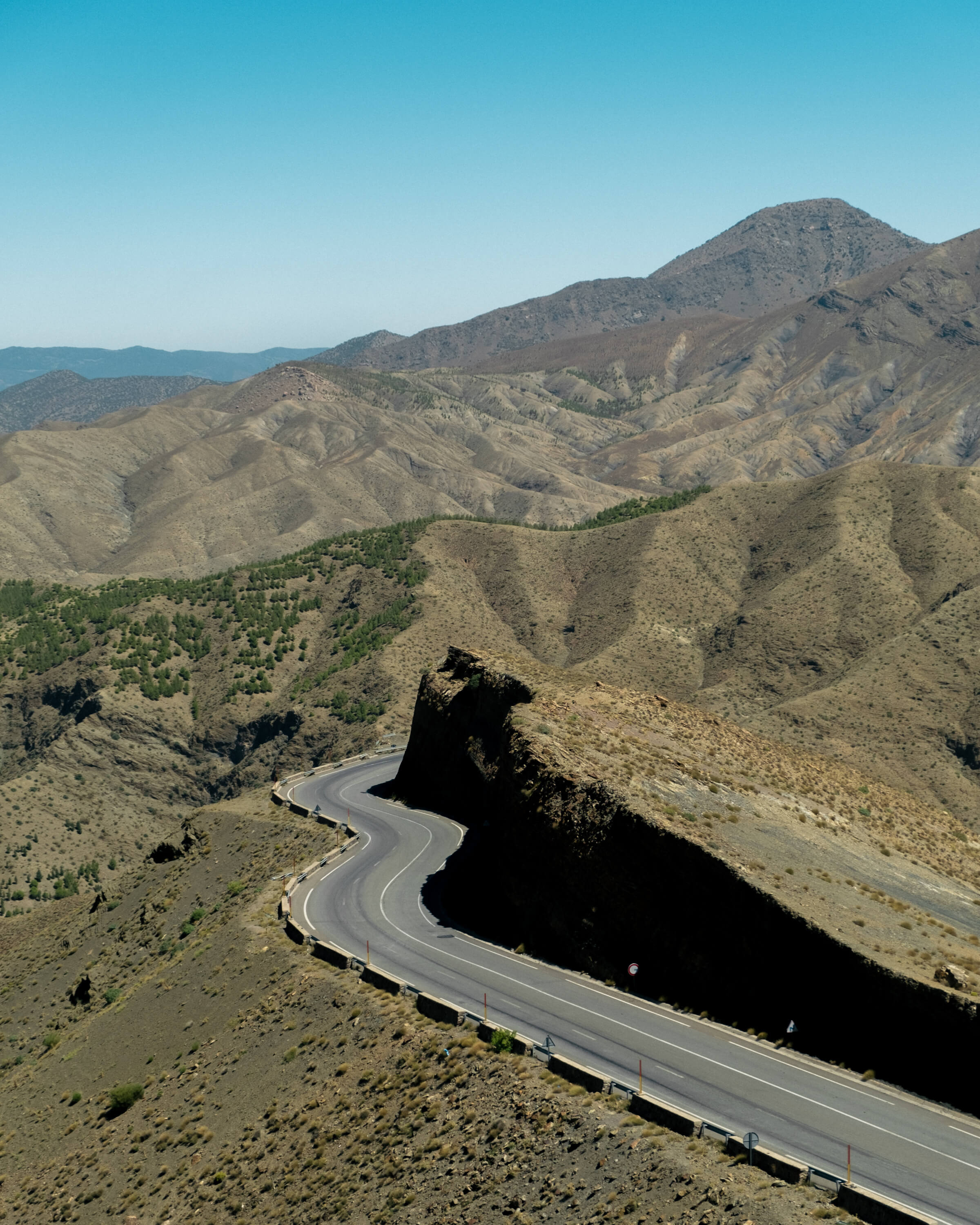 A winding asphalt road curves sharply through the rugged Tizi n'Tichka pass in the High Atlas Mountains of Morocco, surrounded by arid, rocky peaks under a clear blue sky, during a guided tour with Moroccan Guides Travel.