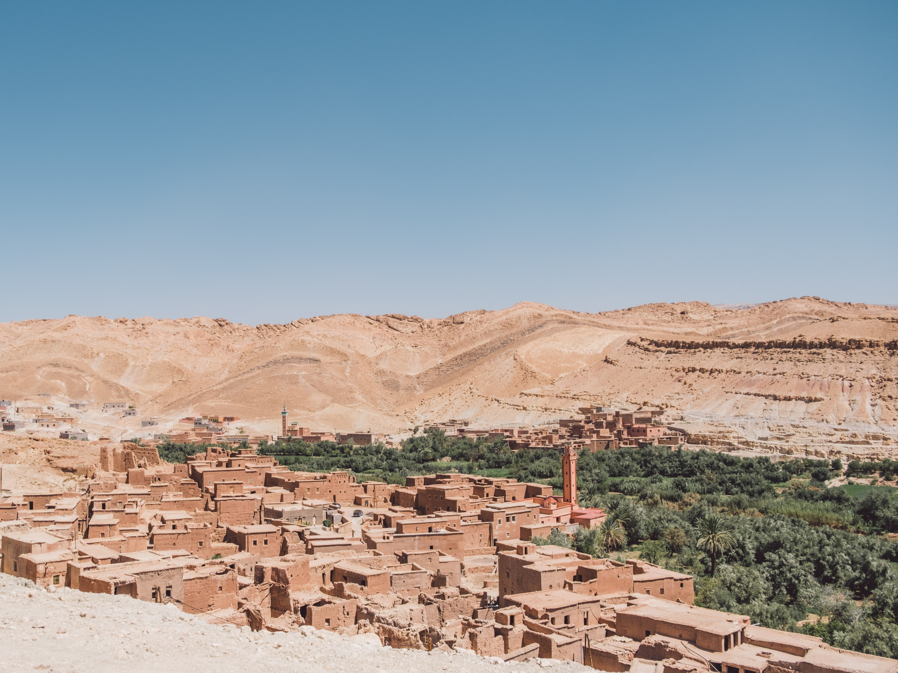 A panoramic view of the red-earthen architecture of a village in Morocco, likely Ait Benhaddou, nestled against a steep, dry, ochre-colored hillside. A line of lush green palm and olive trees separates the buildings from the arid slope, and a mosque