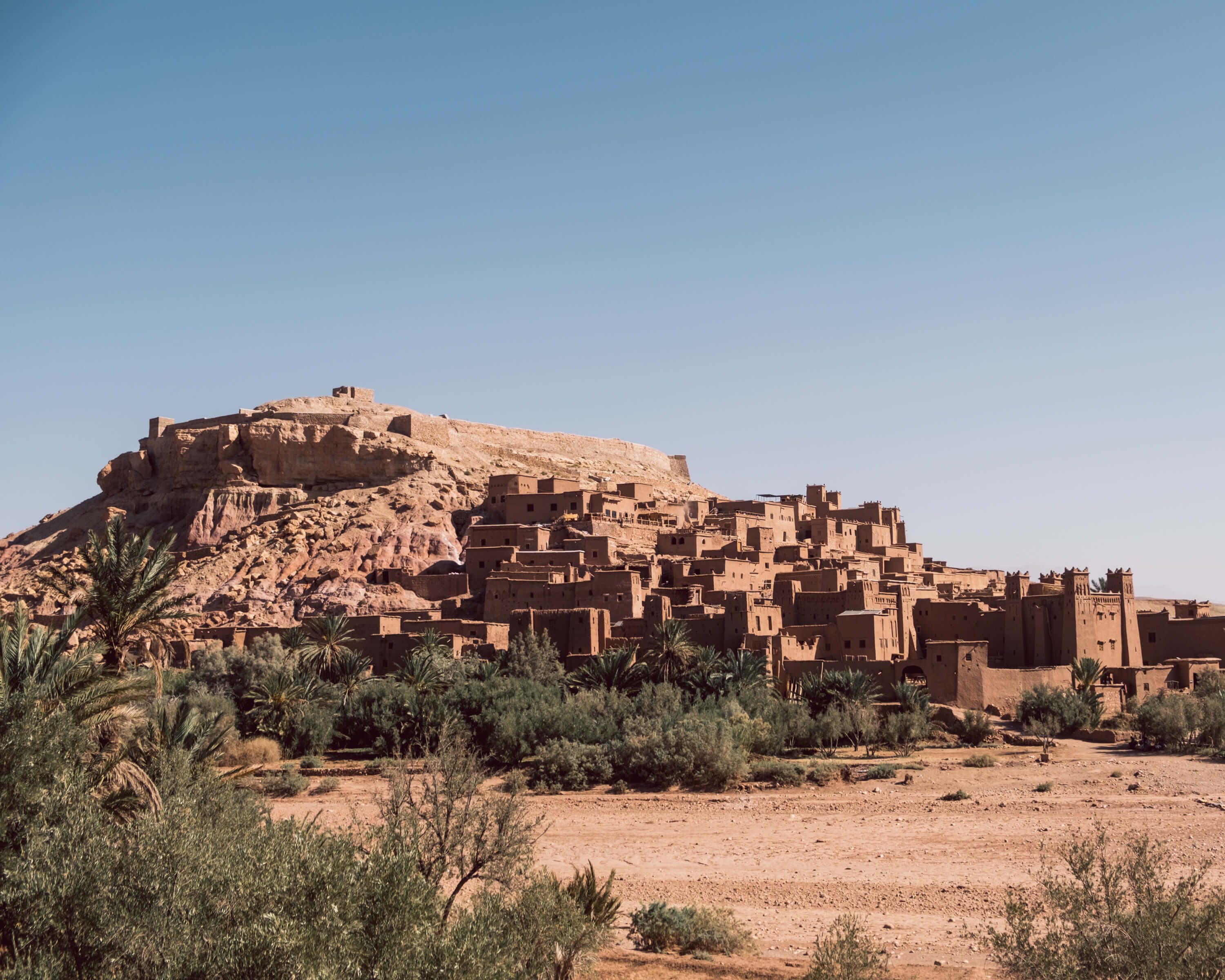 A wide, sunny view of the fortified mud-brick village (ksar) of Ait Benhaddou, Morocco, built against a large, dry, ochre-colored hill. A band of lush green palm trees and foliage separates the cluster of traditional red-earthen buildings.