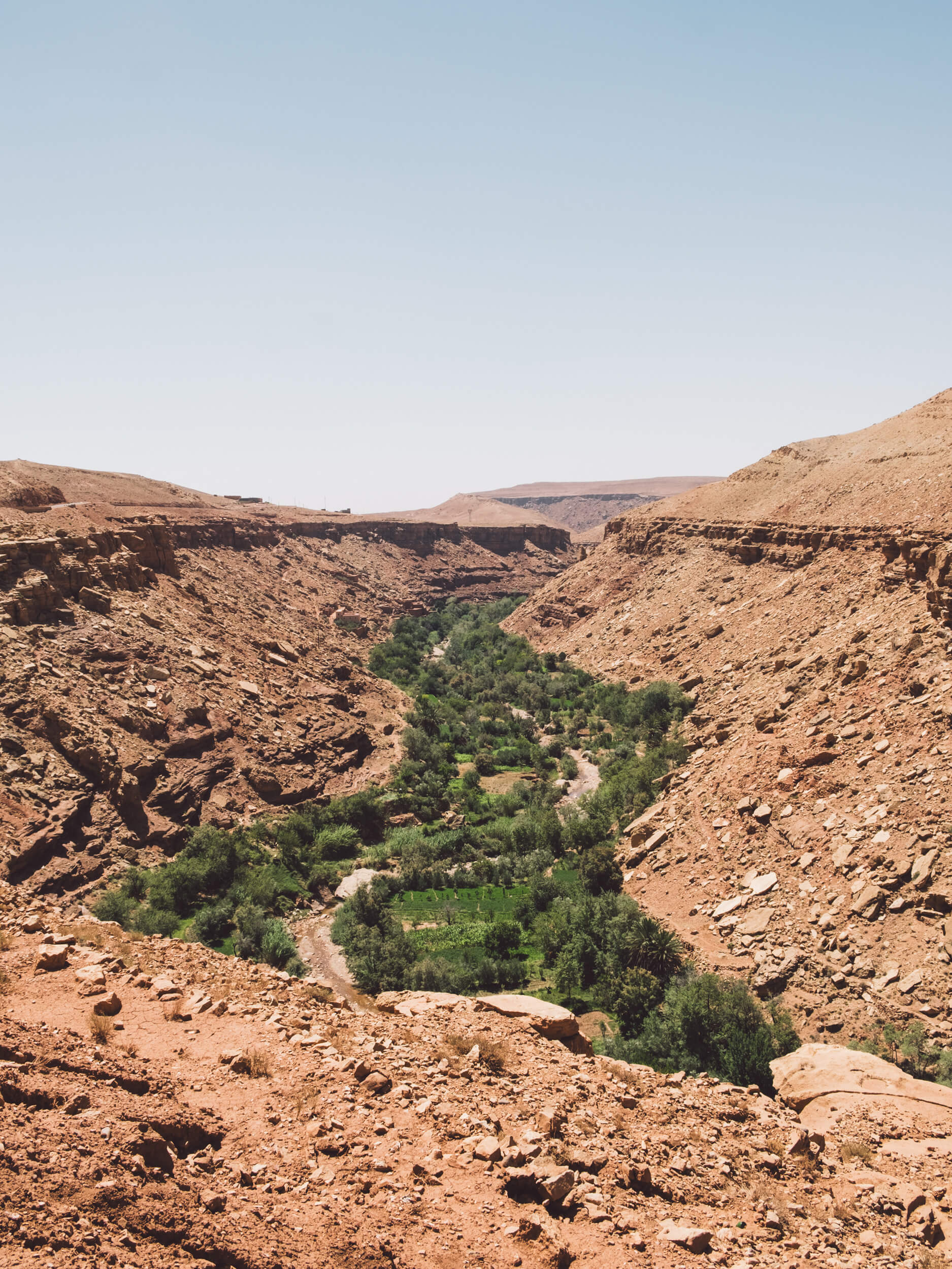A wide vertical shot looking down into a deep, arid valley or gorge, likely in a Moroccan desert region. The slopes of the canyon are steep, rocky, and colored in shades of red and brown.