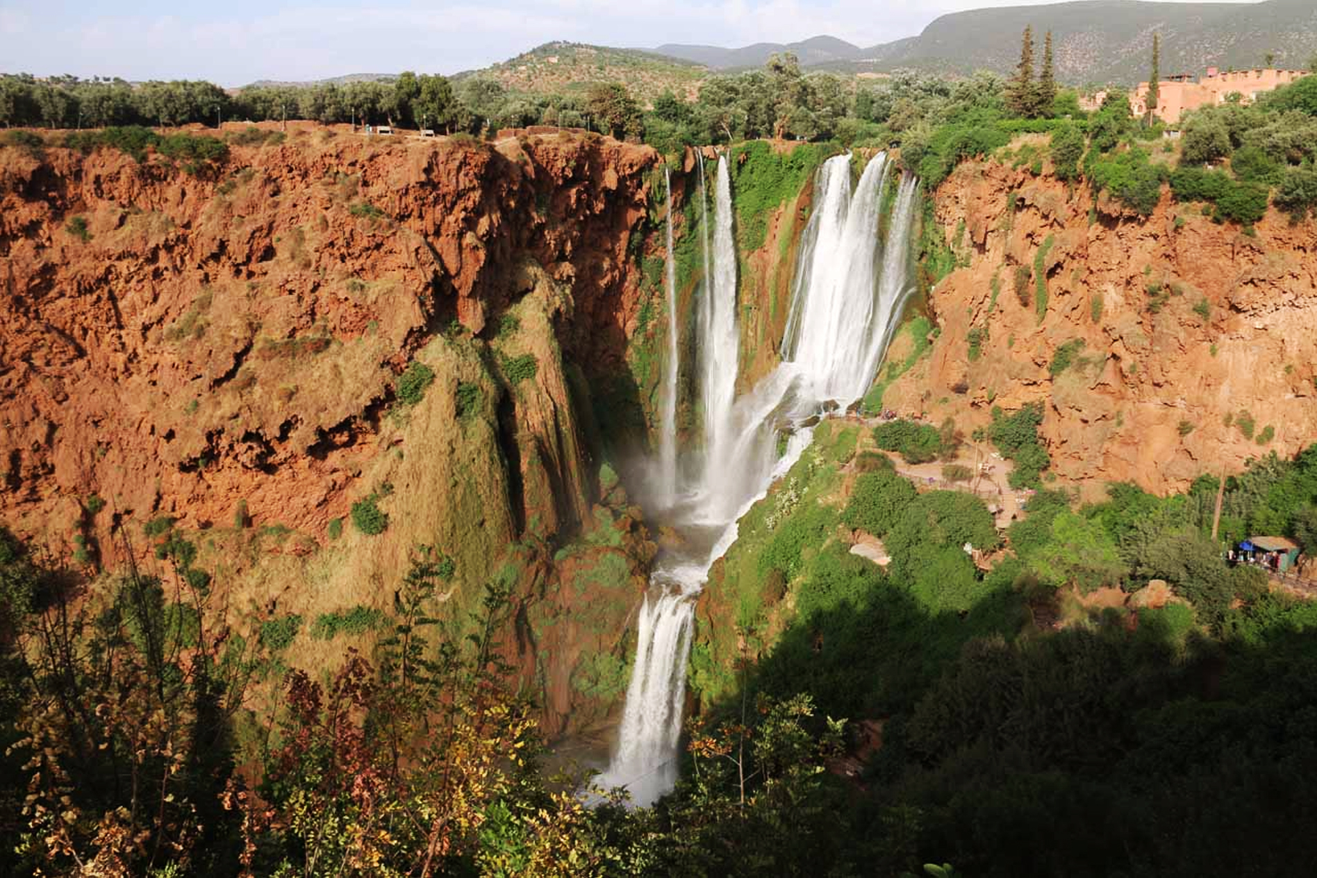 Moroccan Guides Travel presents the magnificent Ouzoud Waterfalls in Morocco, cascading in multiple white streams over sheer, rust-red cliffs and surrounded by thick green vegetation. The view shows the main drop of the falls from above the gorge.
