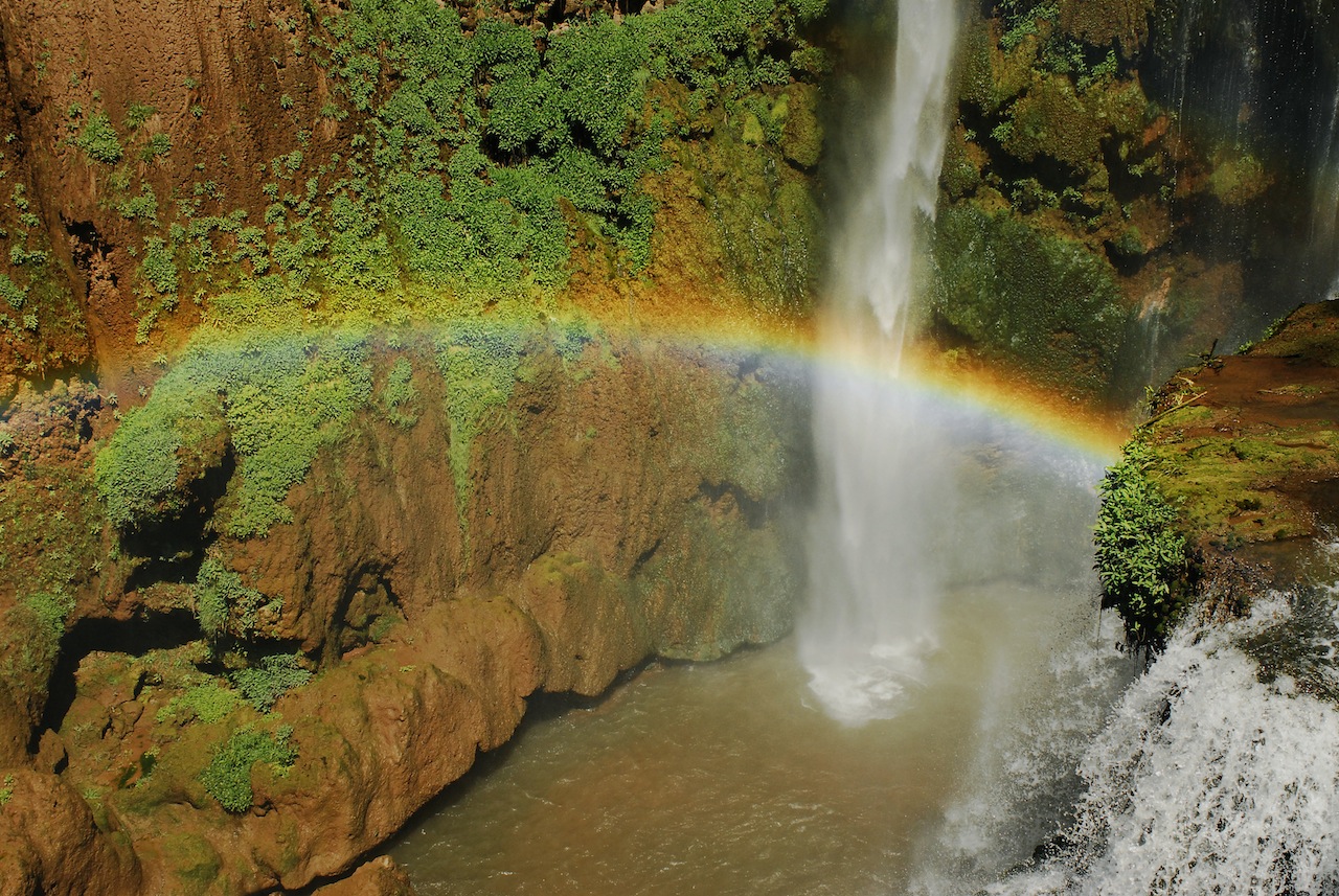 Moroccan Guides Travel showcases a close-up view of the Ouzoud Waterfalls with a magnificent rainbow arching through the mist. The single, powerful stream of water plunges down into a pool, surrounded by the wet, reddish-brown cliffs