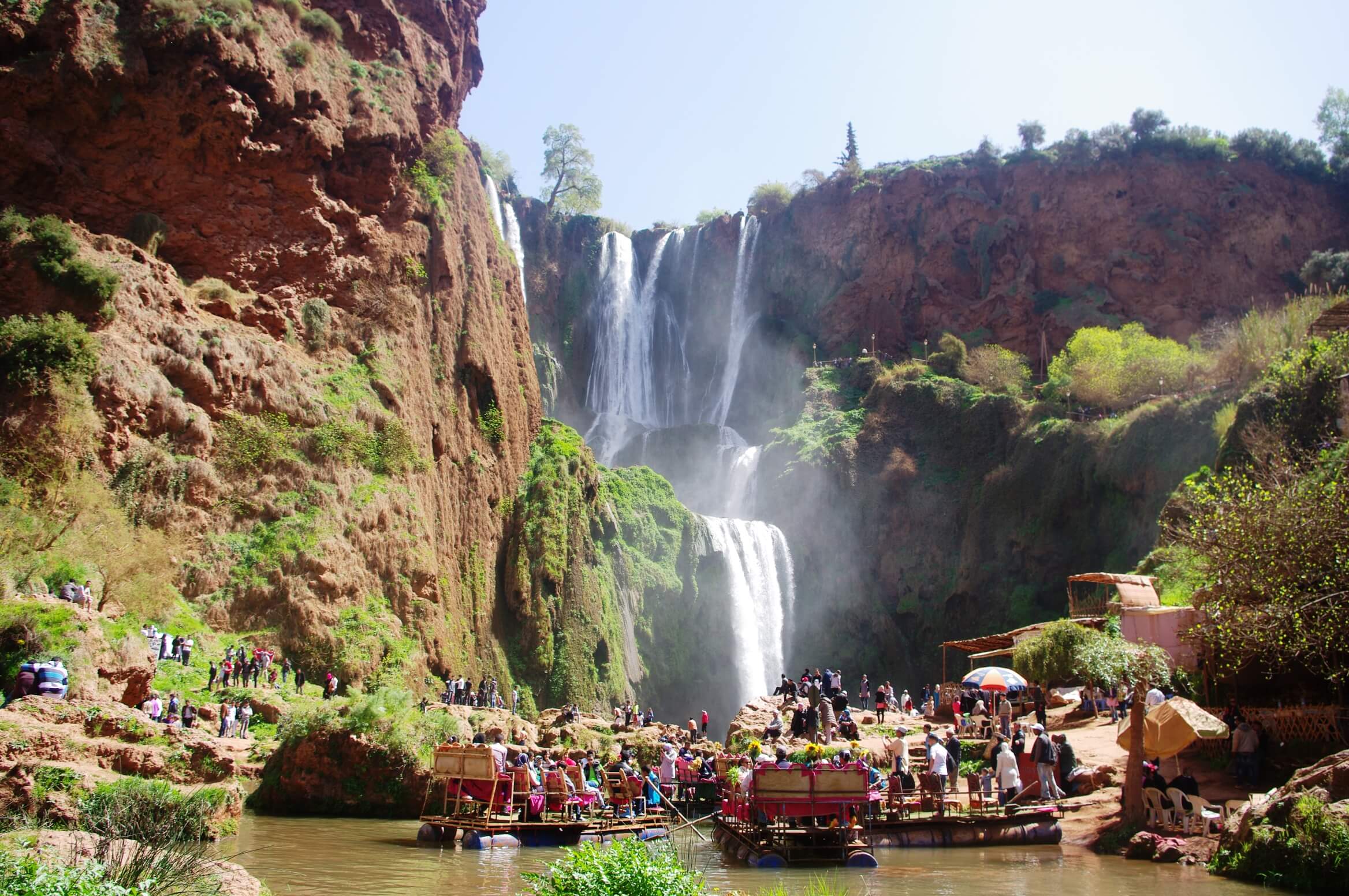 The base of the Ouzoud Waterfalls in Morocco. The multi-tiered cascades plunge over the red-rock cliffs into a large pool where many visitors are enjoying a break on colorfully decorated, rustic rafts.