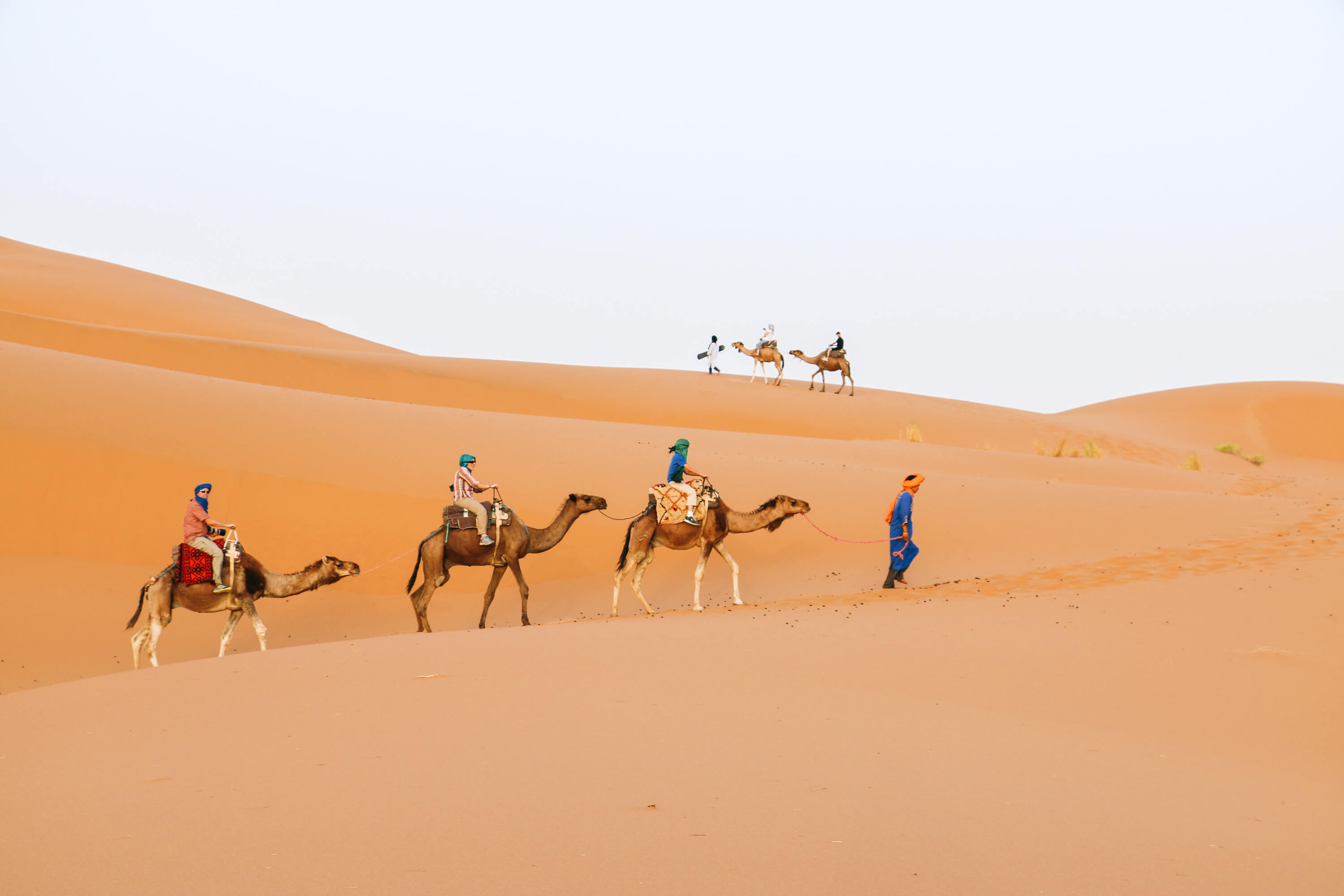 Camel caravan with riders walking across large orange sand dunes in Moroccan desert.
