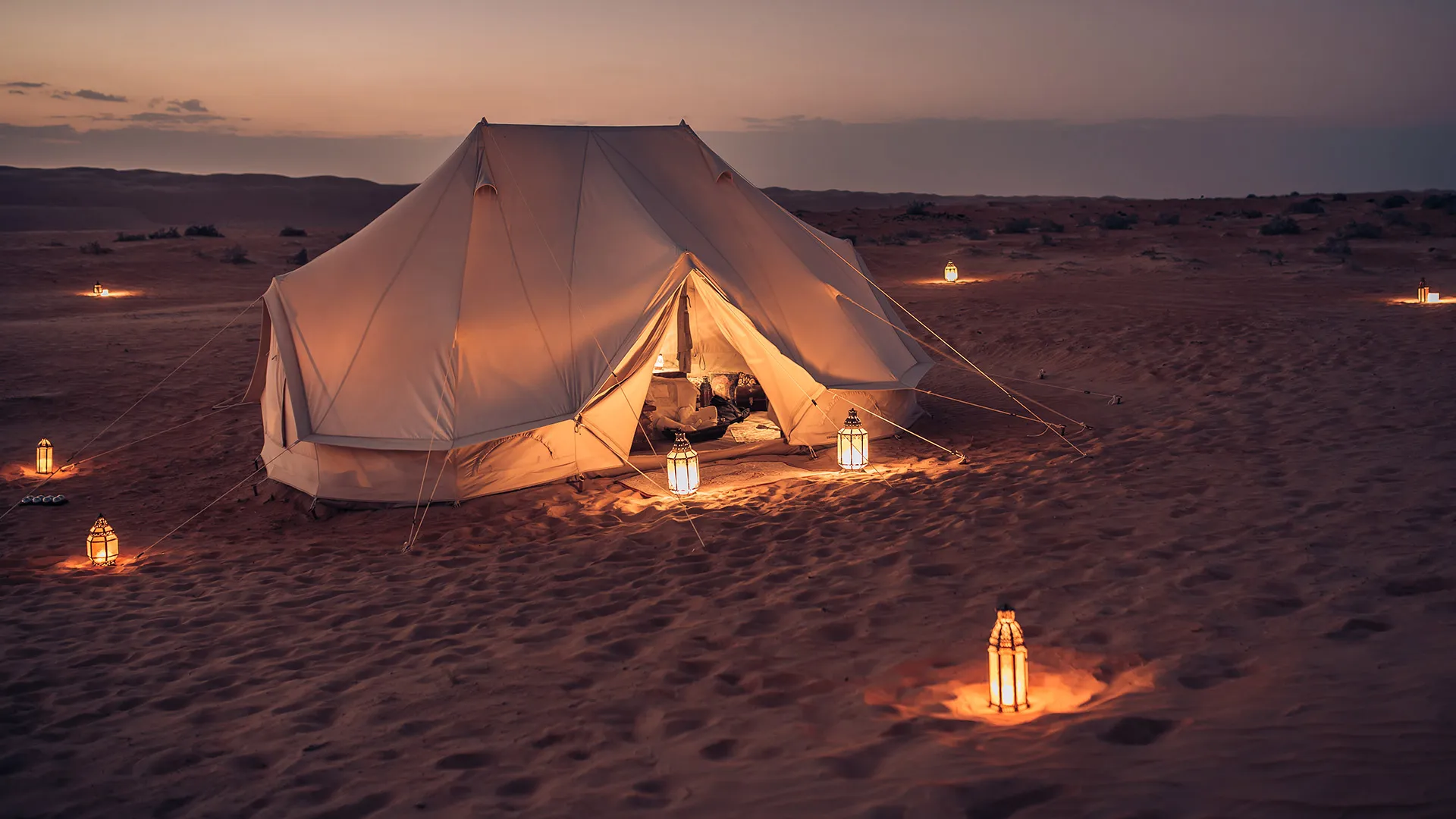 Glowing lanterns illuminate a white tent in a serene Agafay desert landscape for dinner experience at dusk