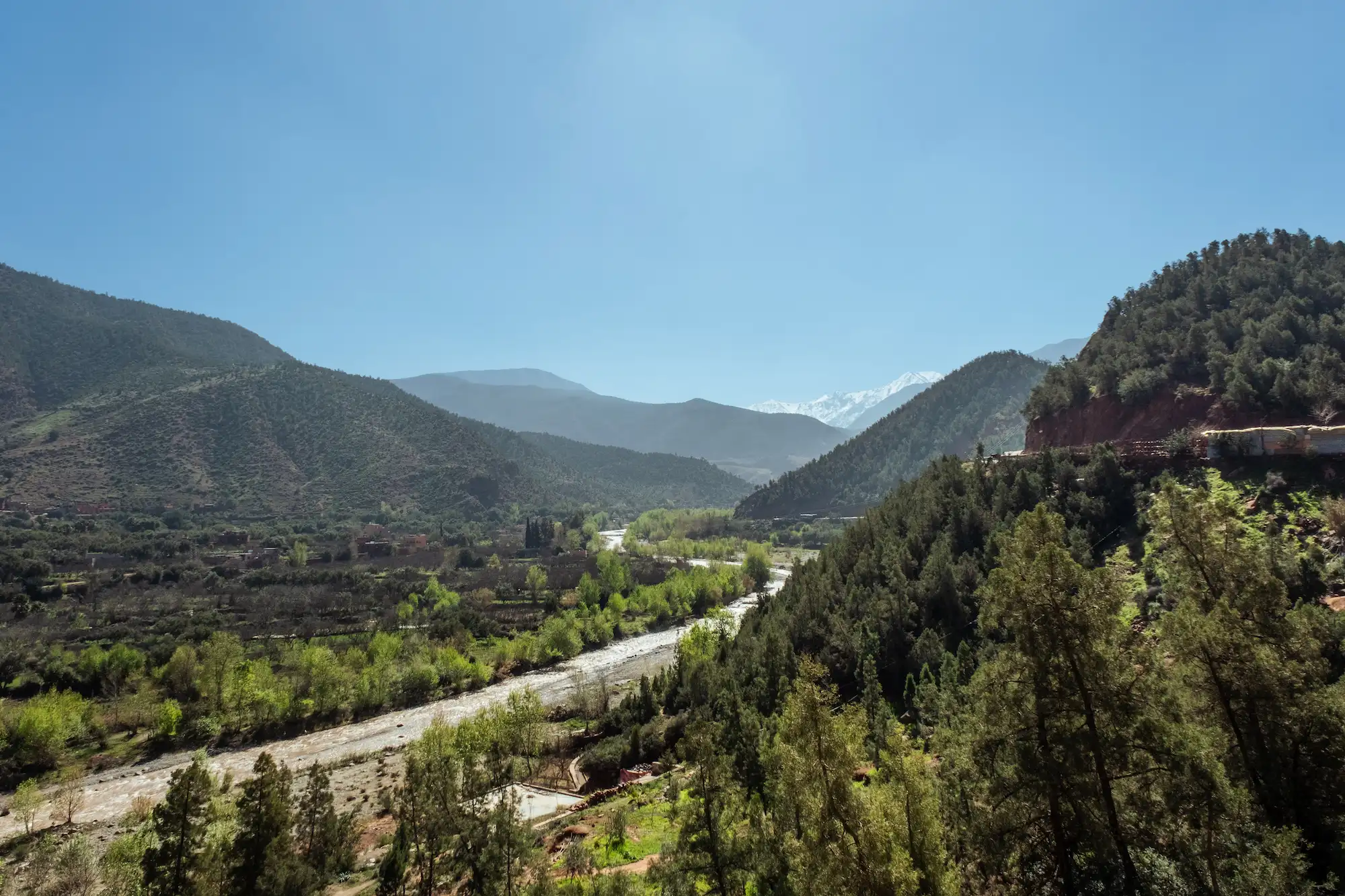 Mountain Ourika valley with winding river, lush trees, and distant snow-capped peaks during a trip with Moroccan Guides Travel.